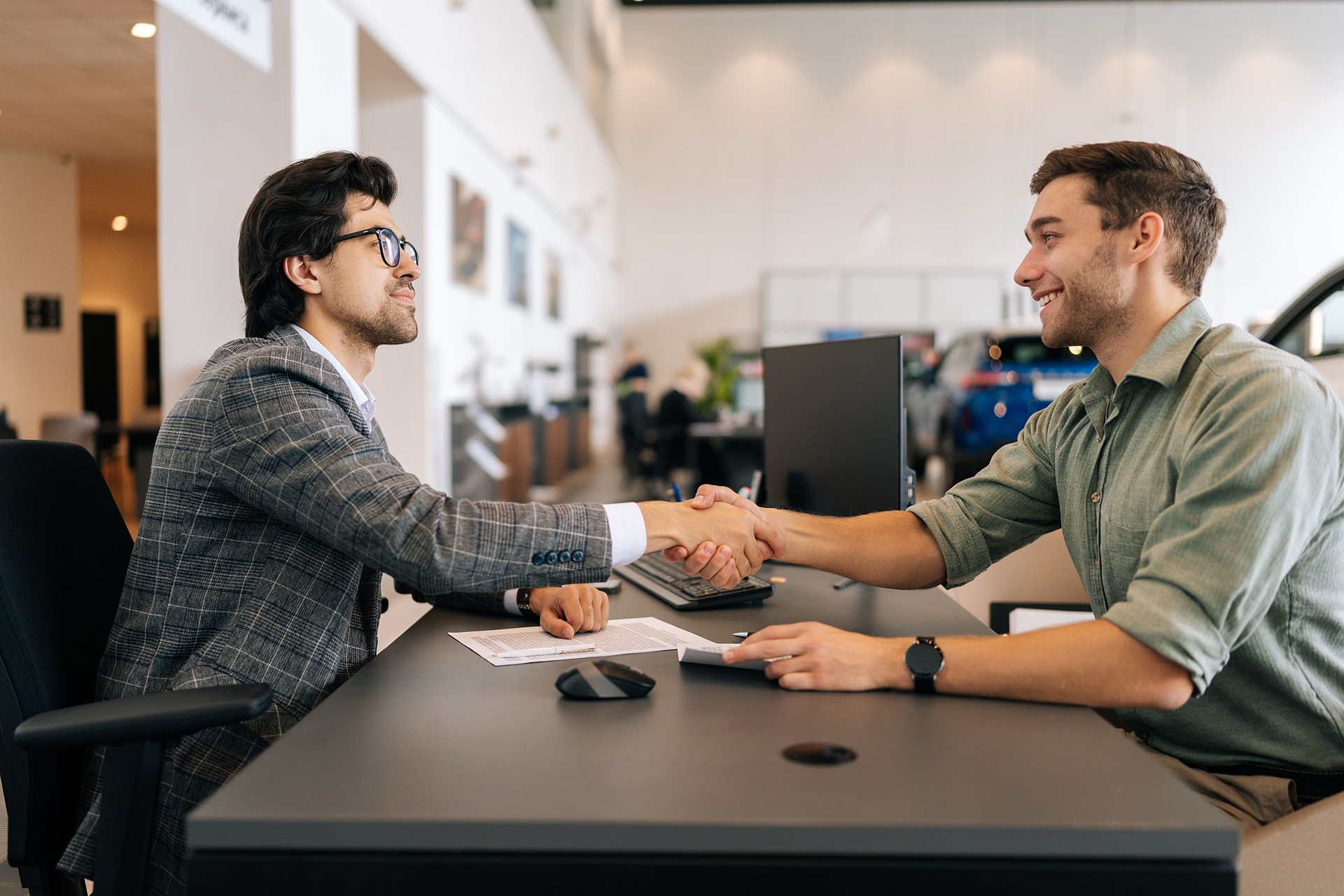 Side view of happy smiling client male purchasing automobile in dealership, signing paper, shaking hands with car dealer in suit enjoying successful agreement.