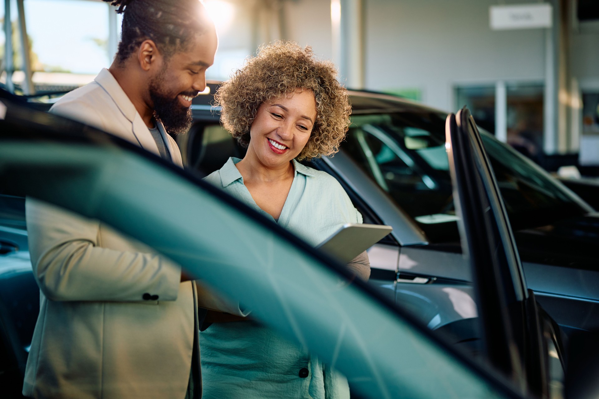 Happy woman using digital tablet with a salesman while buying new car in showroom.